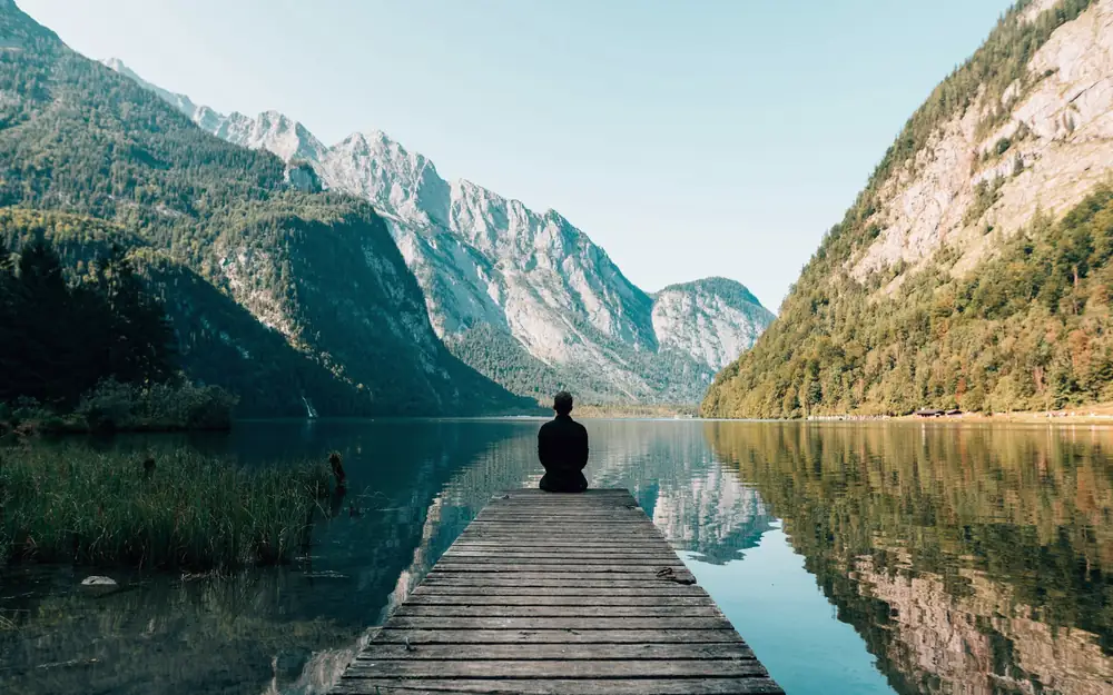 A person sitting at the end of a wooden dock on a perfectly still mountain lake