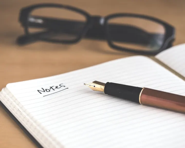An open notebook with a fountain pen and reading glasses on a wooden desk