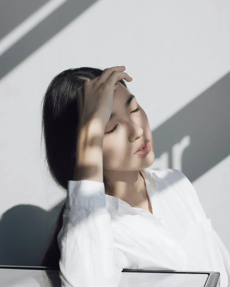 A woman resting her hand on her forehead with her eyes closed in soft window light